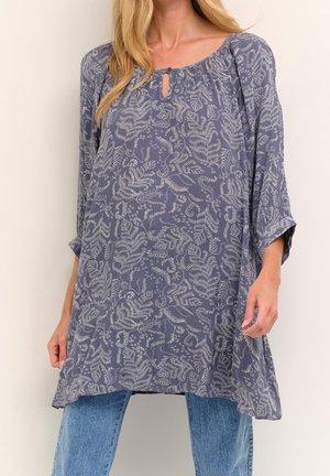 Femme portant une tunique grise ample à manches longues avec des motifs de feuilles blanches, associée à un jean bleu clair sur fond uni.