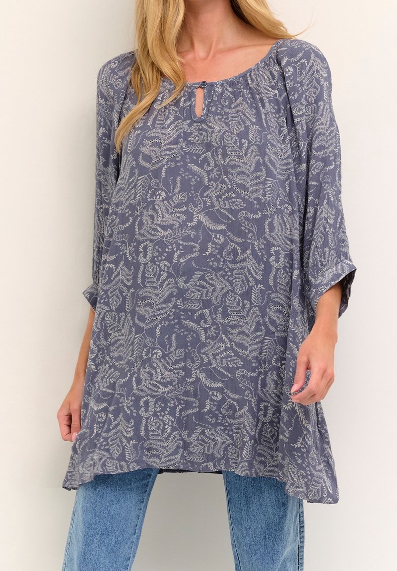 Femme portant une tunique grise ample à manches longues avec des motifs de feuilles blanches, associée à un jean bleu clair sur fond uni.