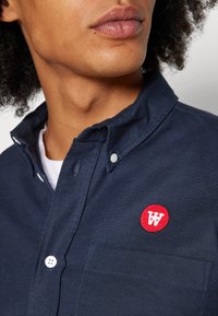 Navy button-up shirt in cotton fabric, featuring a red embroidered logo on the chest pocket and white buttons at the collar and placket.
