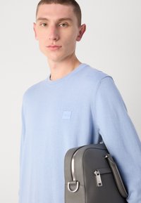 Hellblau gestrickter Pullover mit einem quadratischen Logo-Patch, getragen von einer Person, die eine dunkelgraue Lederhandtasche mit silbernen Beschlägen und einem Reißverschluss hält.