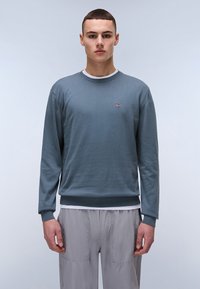 Grijze katoenen trui met een ronde hals, geribbelde manchetten en onderrand, met een klein logo-opdruk. Gelaagd over een wit shirt met zichtbare naden.
