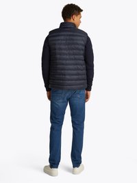 Colete de puffer azul marinho com colarinho alto, textura acolchoada e silhueta ajustada, usado sobre um suéter azul marinho, combinado com jeans azuis e ténis brancos.