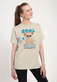 Světle béžové tričko s potiskem karikatury medvěda v slunečních brýlích. Modrý a černý text zní "VYVINUTO V ČASE" s hravými akcenty.