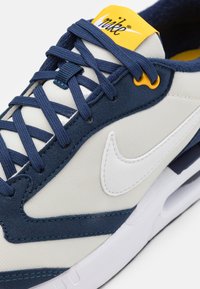 Tenisky Nike s kombinací krémové plátna a námořnického semiše, s bílým logem swoosh, námořnickými tkaničkami, žlutým jazykem a bílou podrážkou.