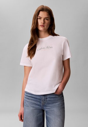 Młoda kobieta z długimi brązowymi włosami nosi białą koszulkę Calvin Klein i niebieskie jeansy, stoi na jednolitym szarym tle.
