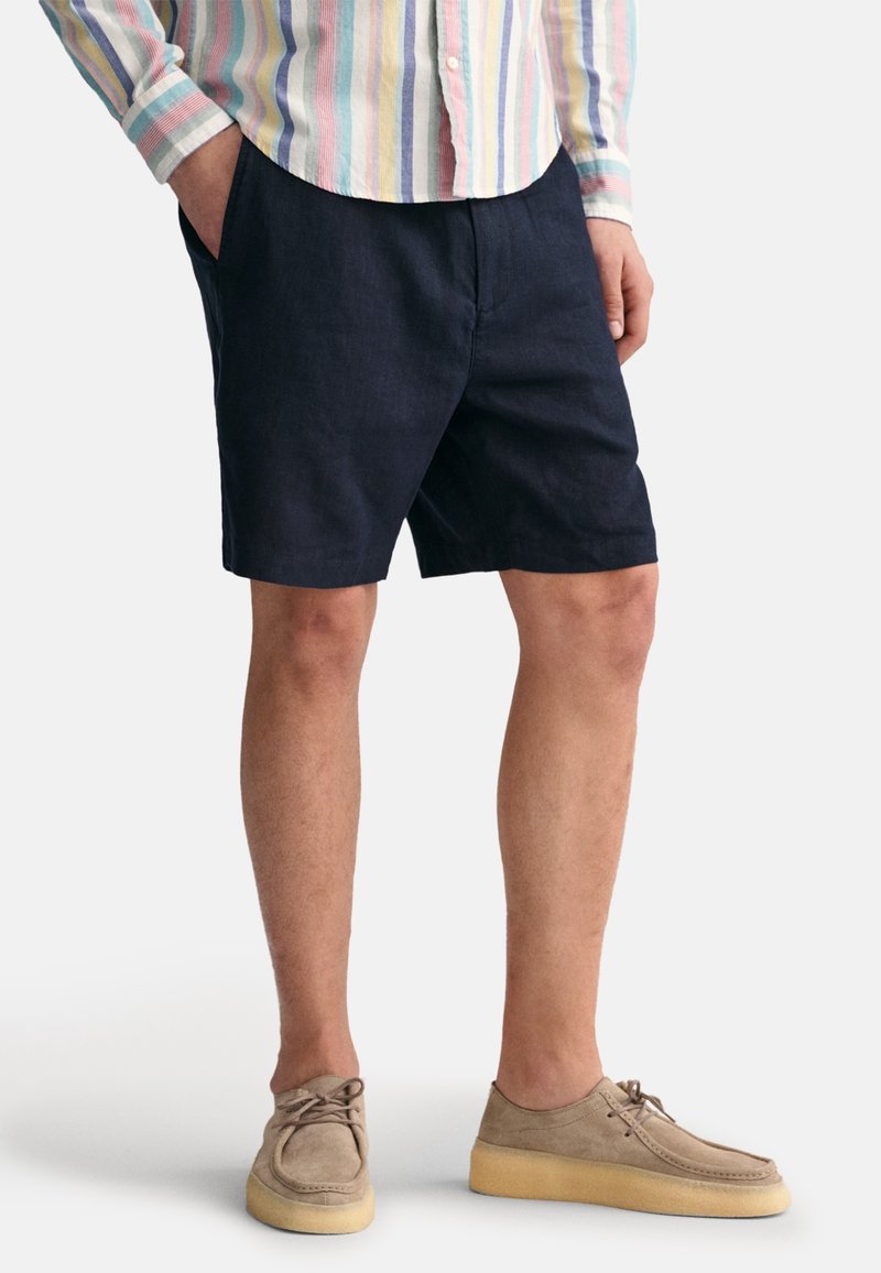 Shorts en lin bleu foncé avec poches latérales, assortis à des chaussures en daim beige clair avec bout rond et semelle en caoutchouc. Chemise rayée multicolore.