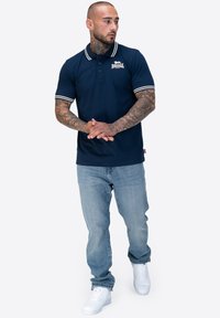 Marineblaues Polohemd mit weiß gestreiftem Kragen und Bündchen, versehen mit einem Logo auf der Brust; kombiniert mit hellblauen Jeans und weißen Sneakers.