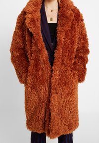 Brun pelsimitation med en shaggy tekstur, åben front og brede revers. Har en løs pasform og en let oversize silhuet.