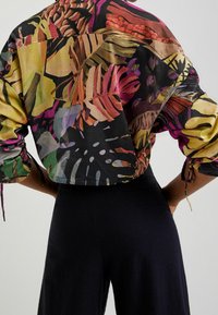 Korte crop shirt met een meerkleurige tropische bladerenprint op een zwarte achtergrond. Driekwartmouwen met strikjes bij de manchetten. Lichtgewicht stof.