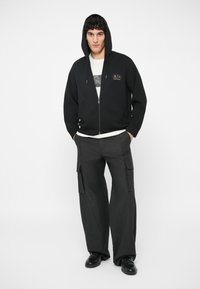 Mladić nosi crnu duksericu s kapuljačom i zipom, bijelu majicu s grafičkim printom, široke crne cargo hlače i crne cipele, stoji s rukama u džepovima.