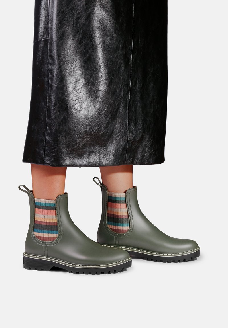 Bottes basses longueur mollet vert olive portées avec des chaussettes rayées multicolores et une jupe en cuir noir brillant sous les genoux.