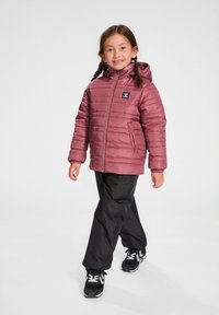 Doudoune violette avec capuche, coutures horizontales et patch avec logo. Portée avec un pantalon noir et des chaussures de sport. Le mannequin est en train de marcher.