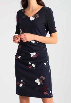 Mujer vestida con un vestido azul marino hasta la rodilla, de manga corta y con un patrón floral en blanco, granate y negro, de pie con las manos entrelazadas.