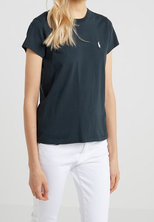 Woman wearing a plain black short-sleeve t-shirt with a small white logo on the chest and white pants against a neutral background.