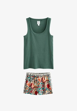 Nő hosszú sötét hajjal, zöld ujjatlan felsőt és színes mintás rövidnadrágot visel, beltéri térben áll egy párnás széknél.