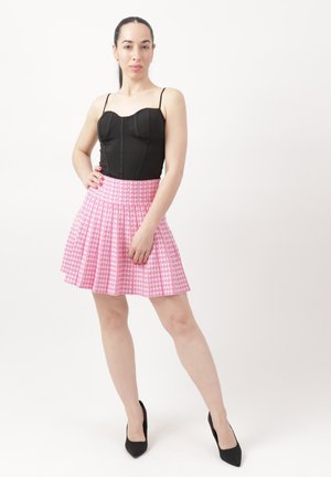 Kobieta stojąca z ręką na biodrze, ubrana w czarny bezrękawnik, różową plisowaną spódnicę w pepitkę i czarne szpilki na białym tle.