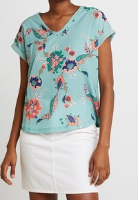 Haut turquoise à manches courtes avec un col en V, arborant des motifs floraux colorés. Associé à une jupe blanche, il a une coupe ample et confortable.