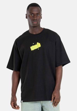 Mann trägt ein übergroßes schwarzes T-Shirt mit kleinem gelbem Motiv in der Mitte, kombiniert mit hellblauen Jeans, steht vor einfachem Hintergrund.