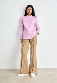 Camicia button-up rosa chiaro a maniche lunghe, abbinata a pantaloni beige a gamba larga e sneakers bianche. Tessuto liscio, design minimalista.