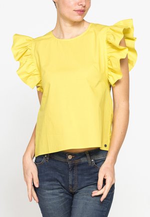 Donna che indossa una maglietta a maniche corte giallo brillante con grandi maniche arricciate e jeans blu scuro, in piedi contro uno sfondo semplice.