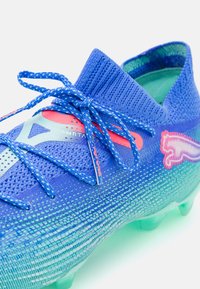 Zapato de fútbol azul con parte superior de tejido texturizado, diseño degradado, acentos rosas y tejido transpirable. Cuenta con cordones llamativos y una suela moldeada.