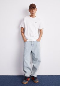 Hvit bomulls T-skjorte med en liten svart "OBEY" logo, lyseblå baggy jeans og grå-svarte sneakers med røde detaljer og gummisåler.