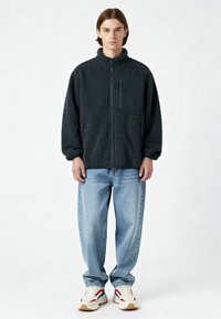 Jeune homme debout, portant une veste polaire noire avec une poche poitrine, un jean bleu clair ample, et des baskets blanches avec des accents rouges et bleus.