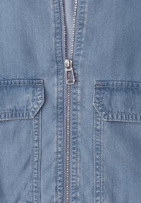 Chaqueta de mezclilla azul claro con cremallera metálica plateada parcialmente cerrada, con dos bolsillos con solapa y costuras visibles.