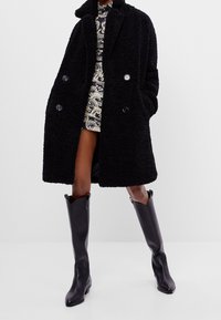 Manteau noir en tissu duveteux à double boutonnage avec de gros boutons et des poches latérales, assorti à des bottes noires élégantes montantes jusqu'au genou, sur un fond uni.