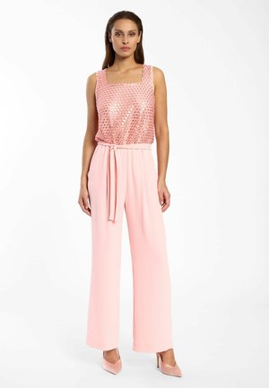 Femme portant un haut rose sans manches orné de sequins et un pantalon rose taille haute à jambes larges avec ceinture, posant devant un fond blanc.