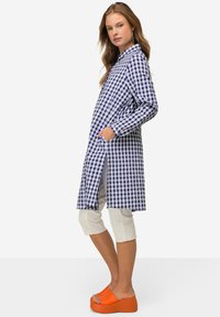 Camicia lunga a scacchi blu e bianca con maniche lunghe, spacchi laterali e due tasche frontali, abbinata a pantaloni capri bianchi e scarpe platform arancioni brillanti.
