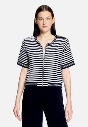 Kvinde med lige brunt hår iført marineblå og hvide vandrette stribede kortærmede zip-up top og sorte bukser, stående mod hvid baggrund.