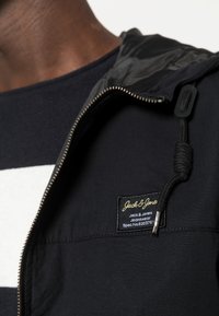 Giacca navy con zip e cappuccio regolabile, con etichetta di tessuto e texture interna a contrasto, abbinata a una camicia a righe sotto.