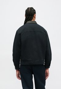 Άτομο με μακριά πλεξούδα γυρνάει την πλάτη, φορώντας μια μαύρη ζακέτα με δερμάτινο γιακά και σκούρα παν trousers σε έναν λευκό φόντο.