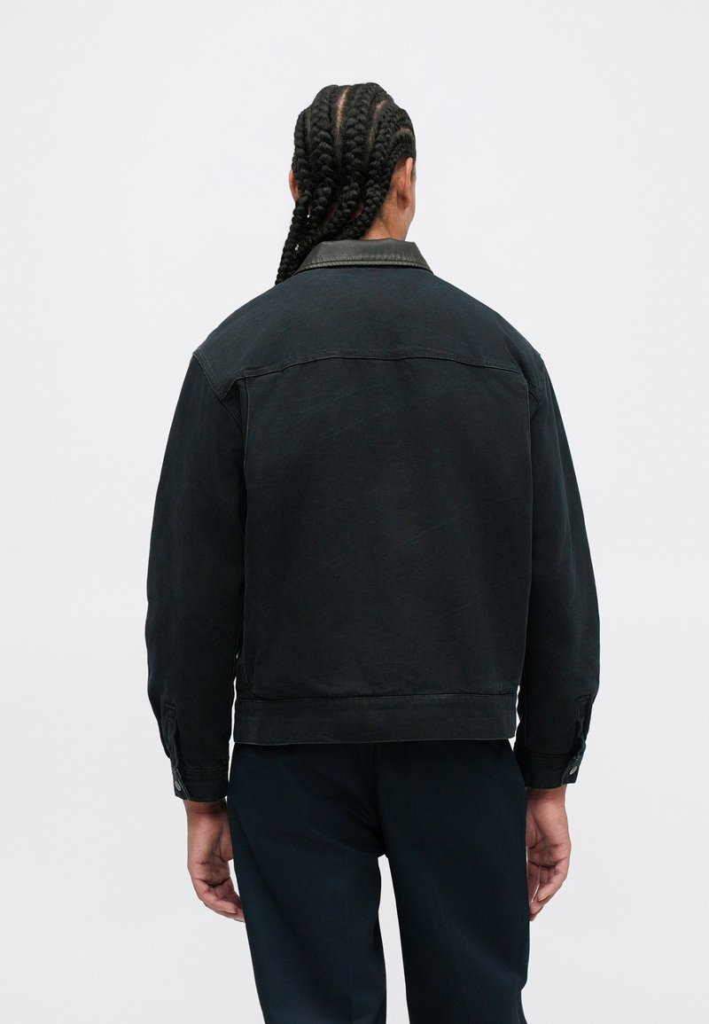 Person with long braided hair stands facing away wearing a black jacket with a leather collar and dark pants against a plain background.