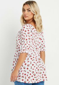 Top floral blanco con pequeñas rosas rojas, mangas abullonadas y un diseño fluido. Tela texturada con suaves rayas. Combinado con jeans azules.