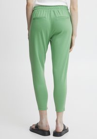 Zelené elastické jogger nohavice so slim fit strihom, s dvomi zadnými vreckami a hladkým materiálom. Nosené s čiernymi plochými sandálmi.