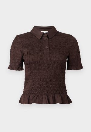 Frau steht mit einer Hand auf der Hüfte, trägt ein braunes kurzärmeliges, strukturiertes Oberteil und hoch taillierte, weiße, weite Hosen vor weißem Hintergrund.