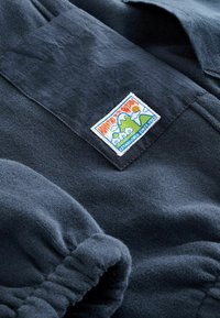 Ciemnoniebieska polarowa odzież z elastycznym mankietem oraz przyszytą prostokątną naszywką przedstawiającą góry, słońce, chmury oraz tekst "Szlak" i "Założono w 1973 roku".