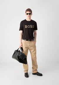 Melnos kokvilnas T-kreklis ar izteiksmīgu "BOSS" logotipu, brūnas chinos bikses, melna ādas rokassoma, saulesbrilles un melna apavi. Minimālistisks dizains.