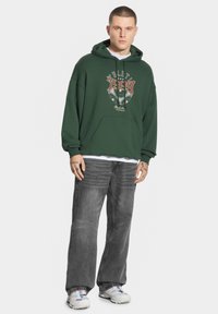 Zielona bluza z kapturem z grafiką, z kieszenią z przodu i luźnym krojem, zestawiona z luźnymi szarymi dżinsami i białymi butami sportowymi.