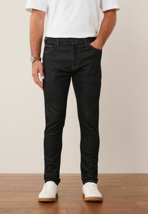 Dark denim jeans with a slim fit, featuring orange stitching, front pockets, and a zip fly, paired with white sneakers and a casual T-shirt.