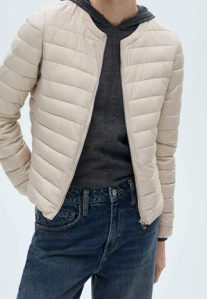 Personne portant une veste matelassée beige clair par-dessus un sweat à capuche gris foncé et un jean bleu foncé, debout les mains dans le dos.
