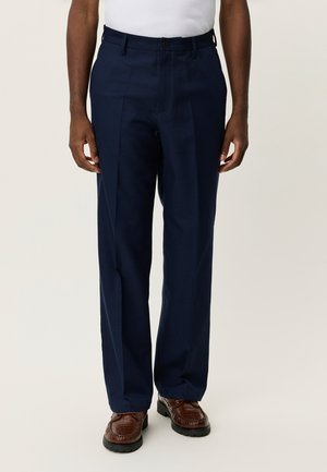 Pantalon habillé bleu marine avec un tissage texturé, devant plissé et poches standards, accompagné de chaussures marron texturées.