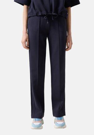 Osoba nosząca granatowe spodnie z plisami z przodu i ściągaczem w talii, zestawione z wielokolorowymi sneakersami w kolorze niebieskim, różowym i białym.