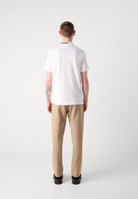 Witte polo met korte mouwen en een zwart geborduurd logo op de kraag, gecombineerd met beige broek en zwarte schoenen.