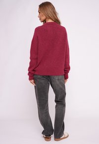 Oversizowy bordowy sweter dziany z prążkowanymi mankietami i ściągaczem, zestawiony z luźnymi szarymi dżinsami i stylizowany z wzorzystymi butami.