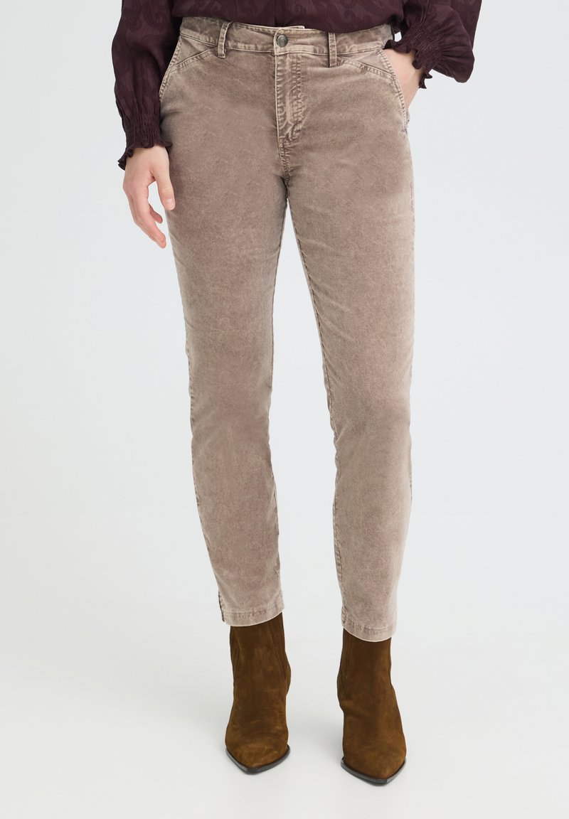 Femme portant un pantalon en velours côtelé taupe taille haute et des bottines en daim marron, une main dans la poche et un chemisier violet foncé.
