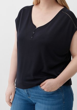 Vrouw draagt een mouwloze zwarte top met knopen en blauwe spijkerbroek, één hand in de zak, tegen een effen lichte achtergrond.