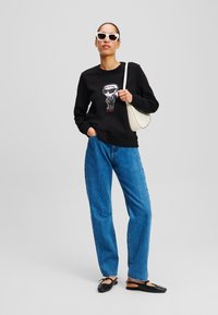 Sudadera negra con diseño gráfico, combinada con jeans de denim azul holgados y sandalias planas negras. Los accesorios incluyen gafas de sol blancas y un bolso crema.